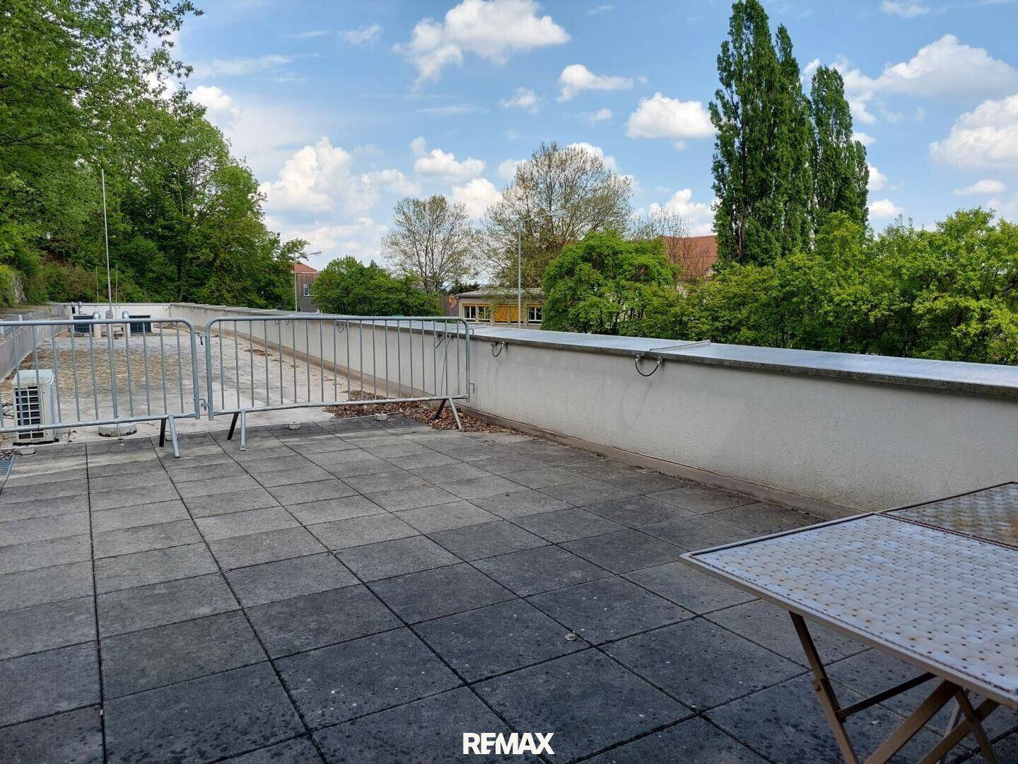 Große Dachterrasse mit Fliesenboden und Metallgeländer, umgeben von Bäumen und Gebäuden.