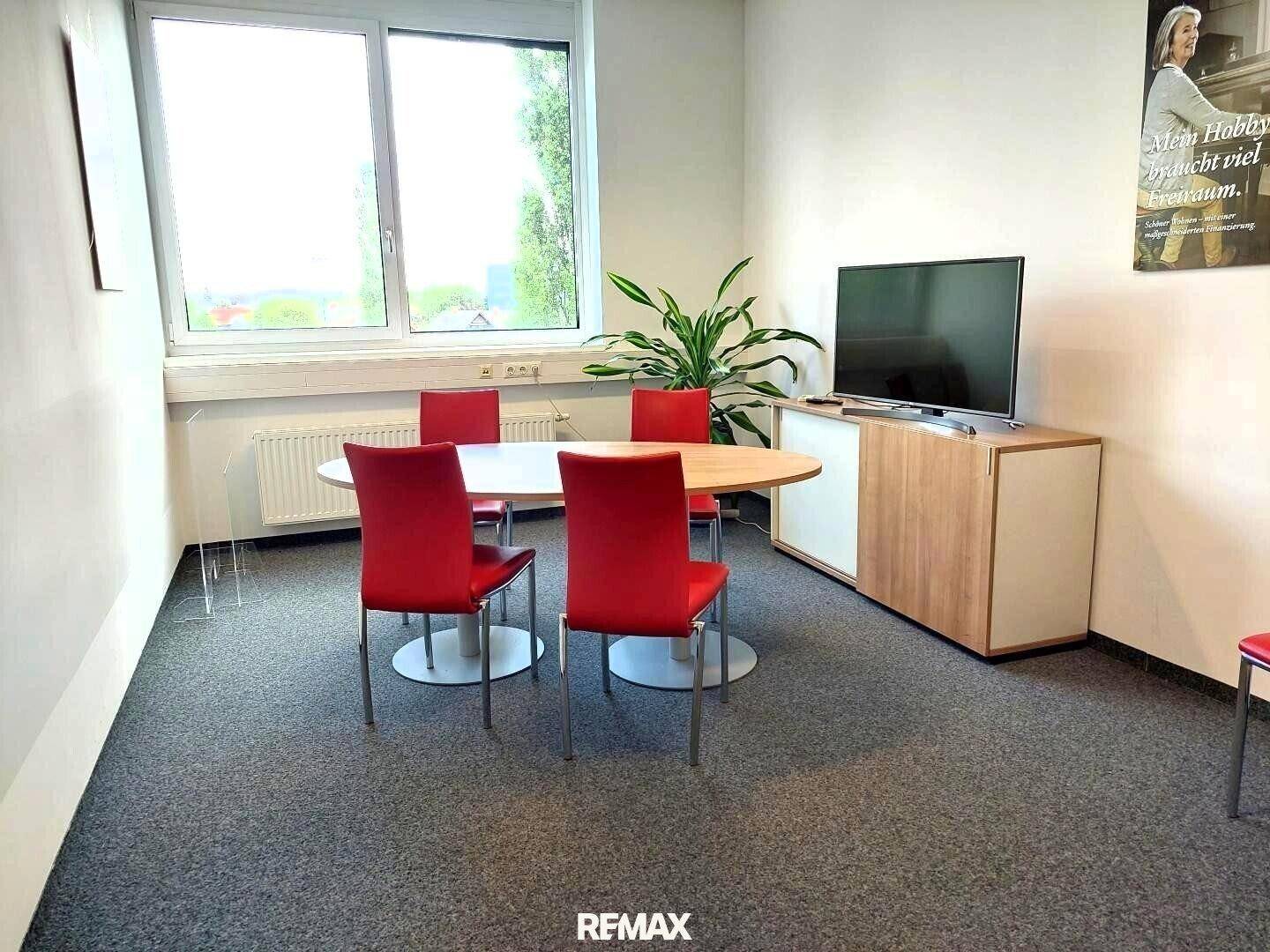 Büro mit Besprechungsecke, Fernseher und großem Fenster, das viel Tageslicht hereinlässt.