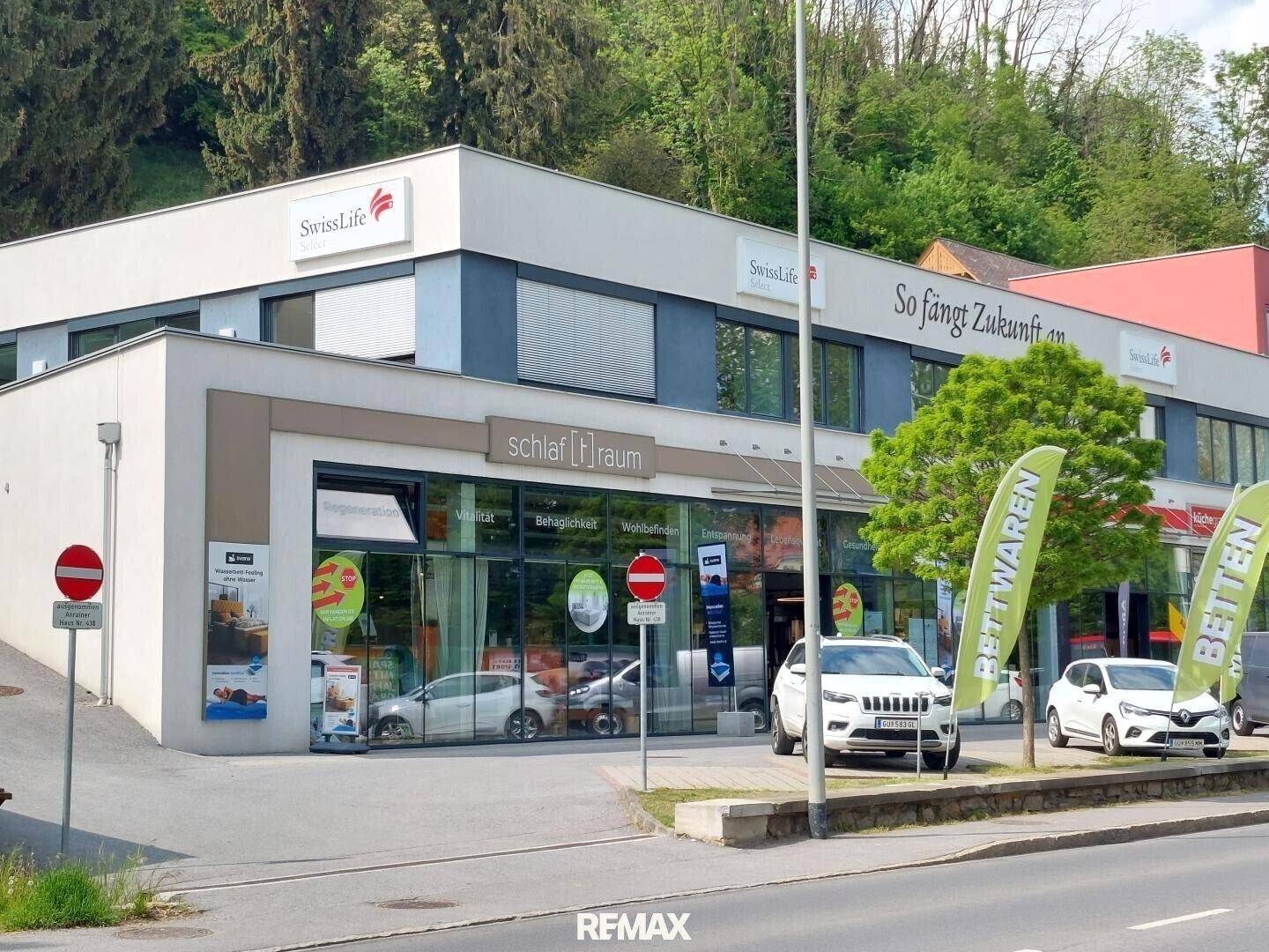 Nahaufnahme des modernen Geschäftsgebäudes mit großen Schaufenstern und Beschilderung, umgeben von Bäumen.