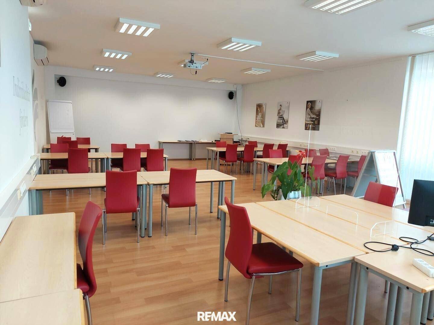 Großer Konferenzraum mit mehreren Tischen und roten Stühlen, ausgestattet mit einem Beamer für Präsentationen.