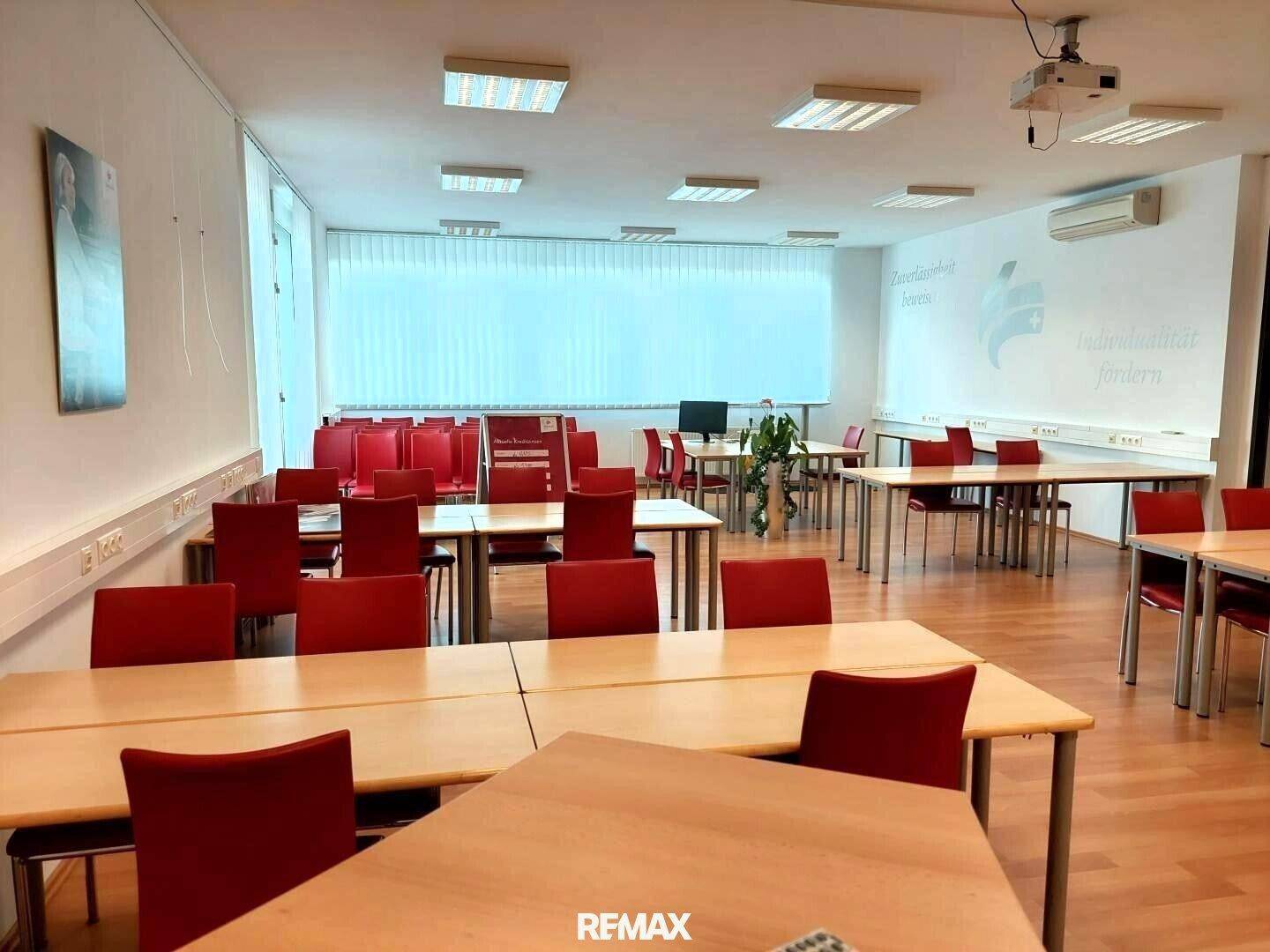 Heller Konferenzraum mit vielen Tischen und roten Stühlen, großen Fenstern und einem Projektor an der Decke.