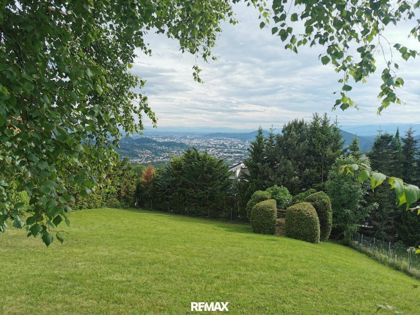 Grünes Grundstück mit Bäumen und einem weiten Blick auf die Stadt unter bewölktem Himmel.
