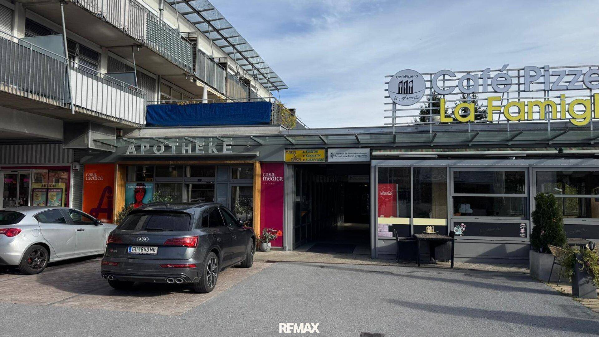 Außenansicht eines Geschäftsgebäudes mit Apotheke, Café und Parkplätzen davor.