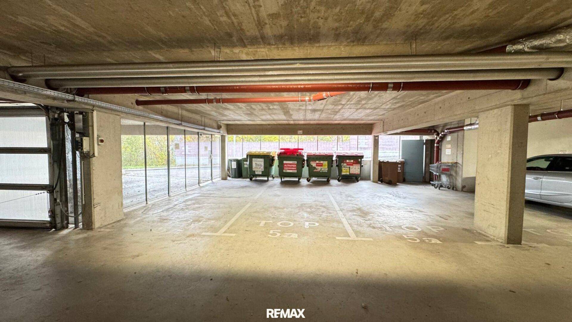 Tiefgarage mit markierten Stellplätzen, Abfallbehältern und sichtbaren Rohren an der Decke.