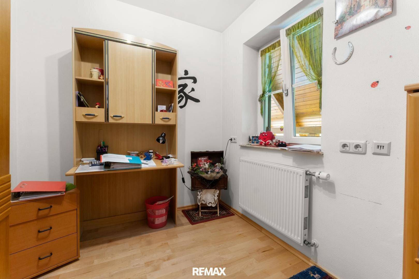 Kleines Arbeitszimmer mit Schreibtisch, Holzschrank und Fenster mit Blick ins Grüne.
