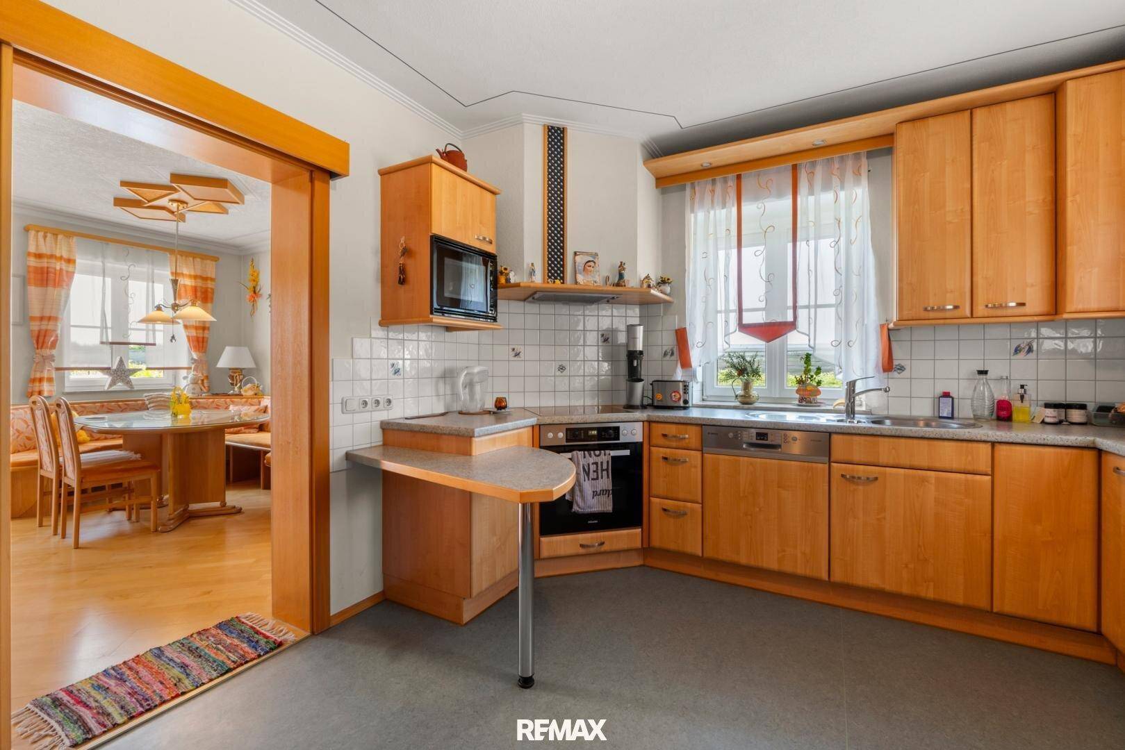 Helle Holzküche mit Einbaugeräten, Fliesenrückwand und Essbereich im Hintergrund.