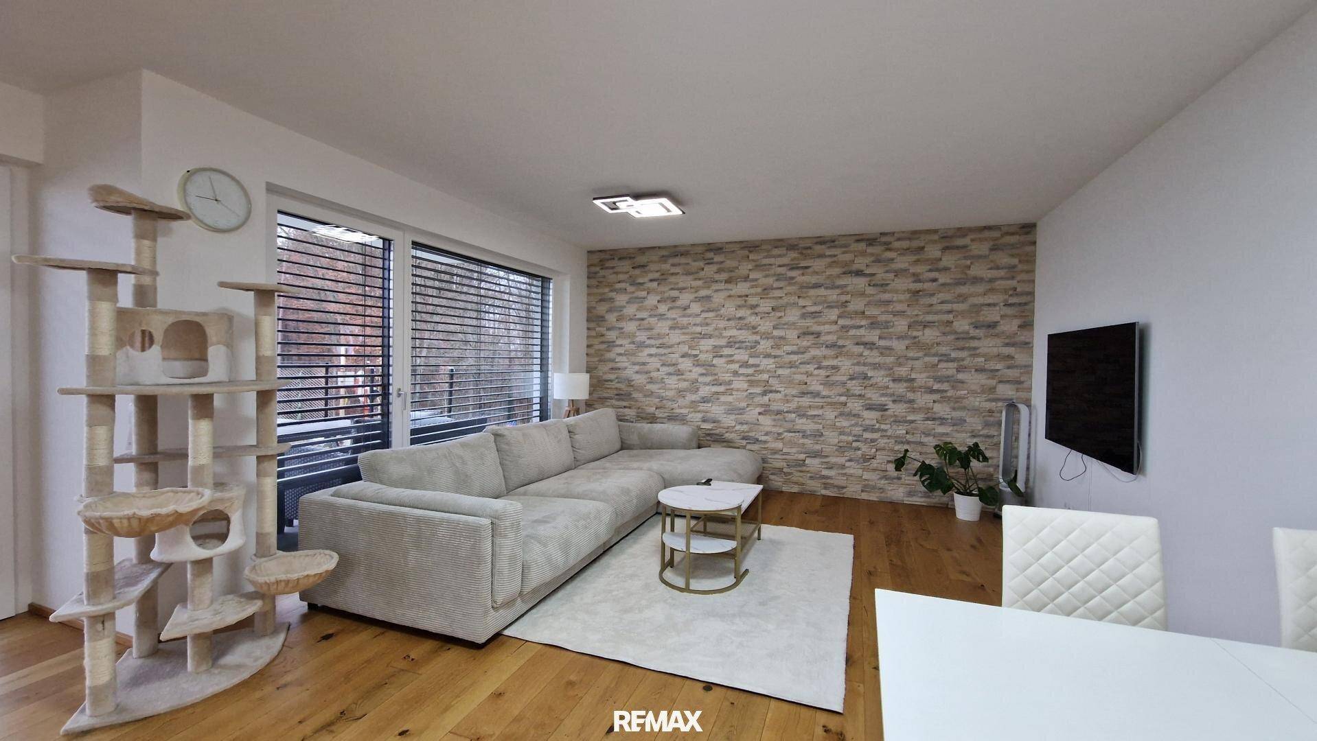 Helles Wohnzimmer mit beigem Sofa, Holzboden, Steinwand und einem Katzenbaum.
