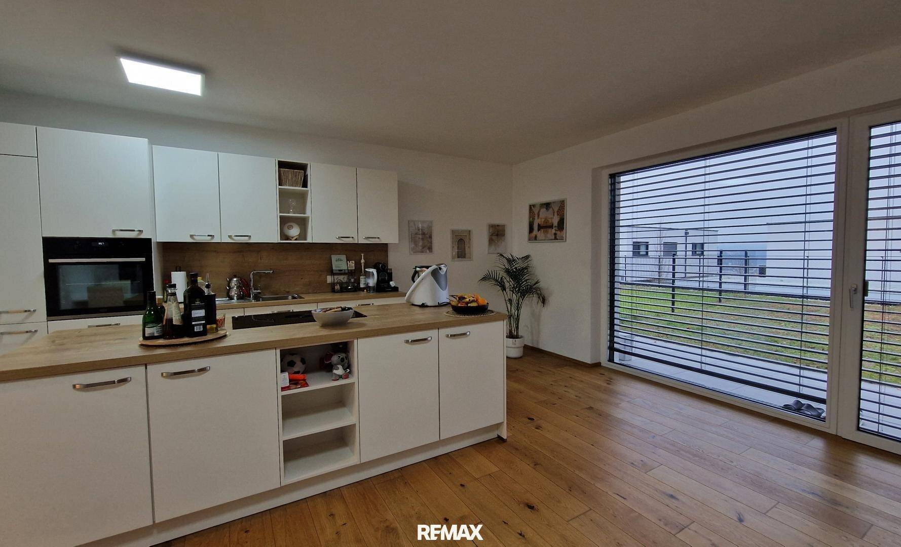 Moderne weiße Küche mit Holzarbeitsplatte, Einbauofen und großem Fenster.