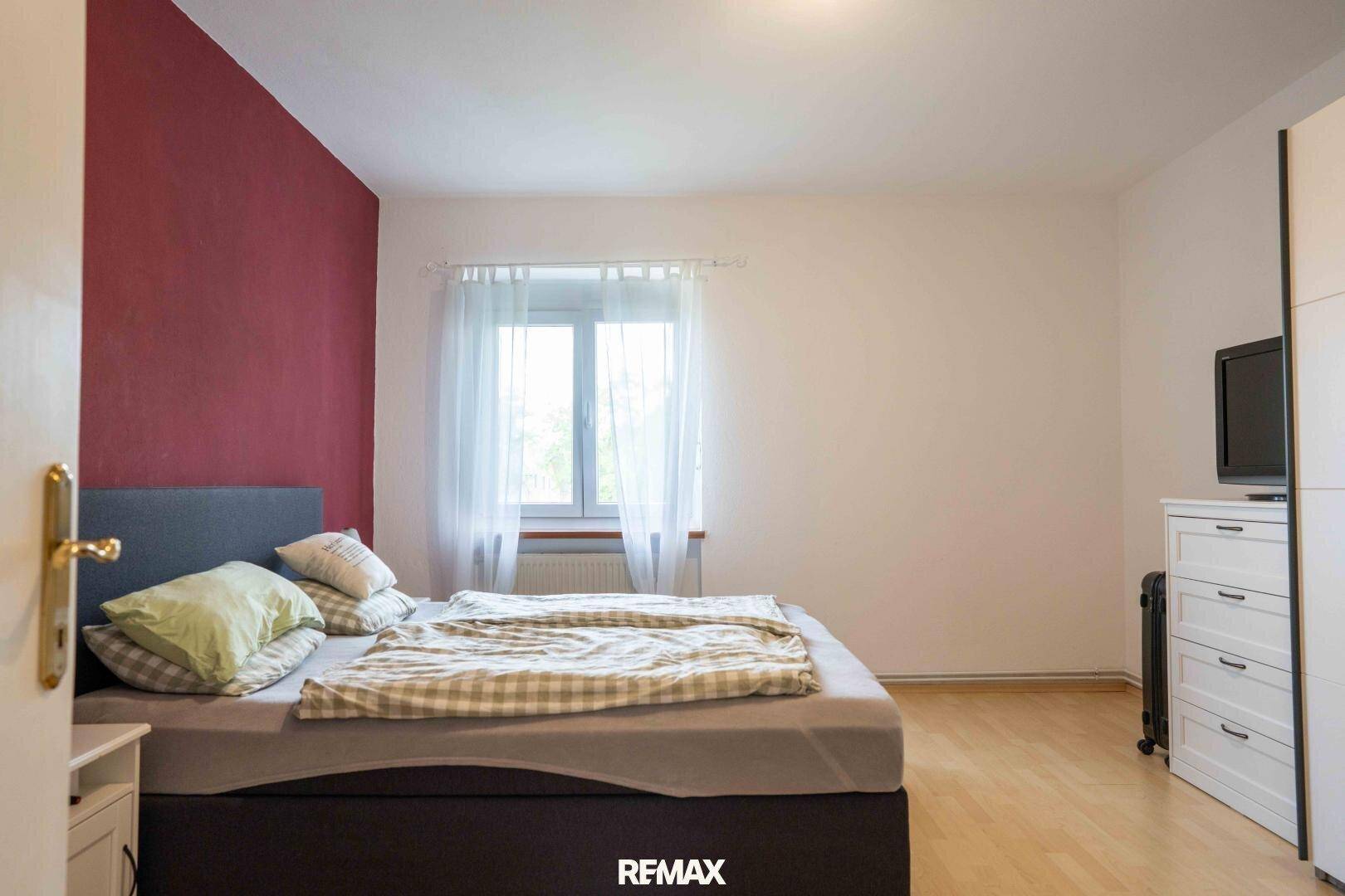 Schlafzimmer mit Doppelbett, roter Akzentwand und hellem Holzboden.