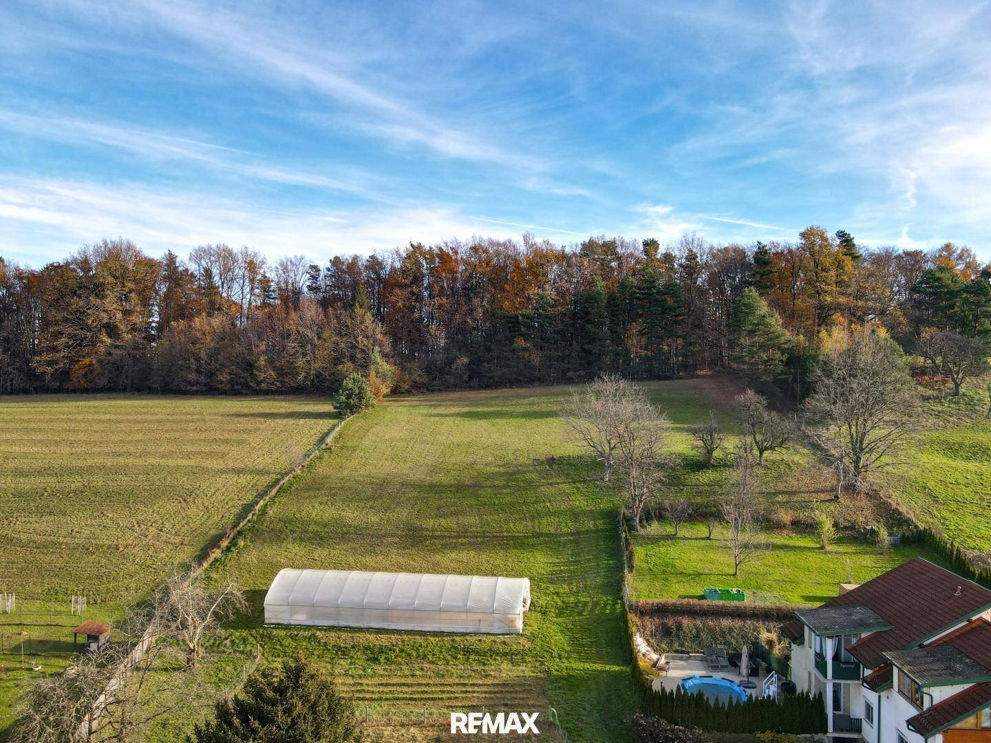 Ländliche Szene mit einem Gewächshaus, einem Haus mit Pool und weiten Feldern im Hintergrund unter blauem Himmel.