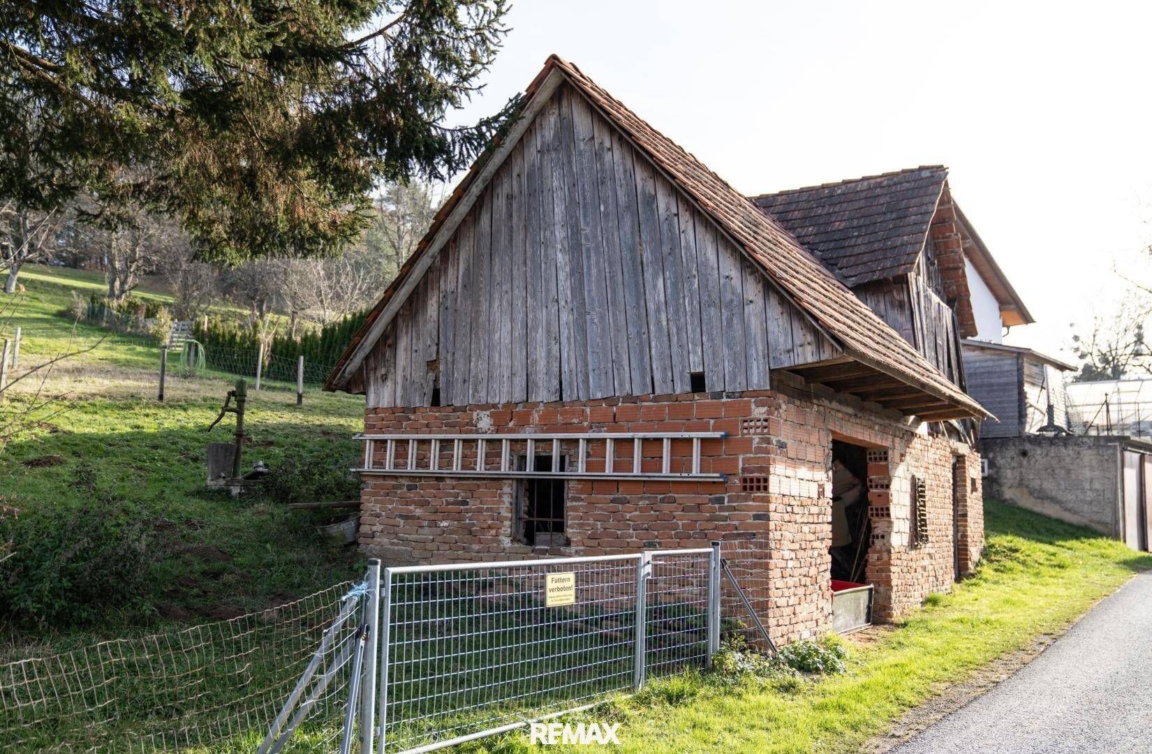 Altes Scheunengebäude aus Ziegeln und Holz mit einem schrägen Dach und einer Leiter, umgeben von Grün.