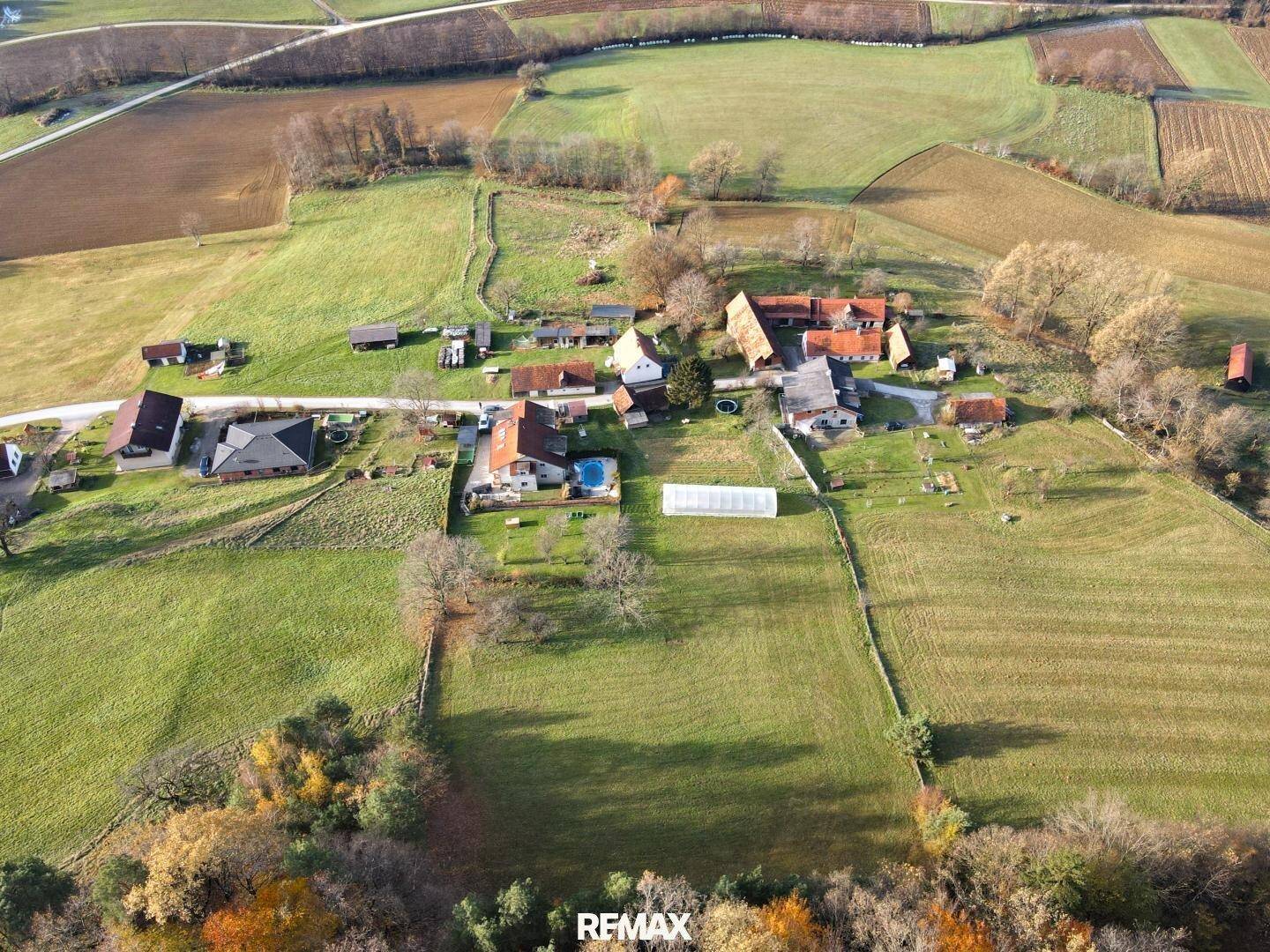 Weitläufige Luftaufnahme einer ländlichen Gegend mit mehreren Häusern und landwirtschaftlichen Flächen unter blauem Himmel.