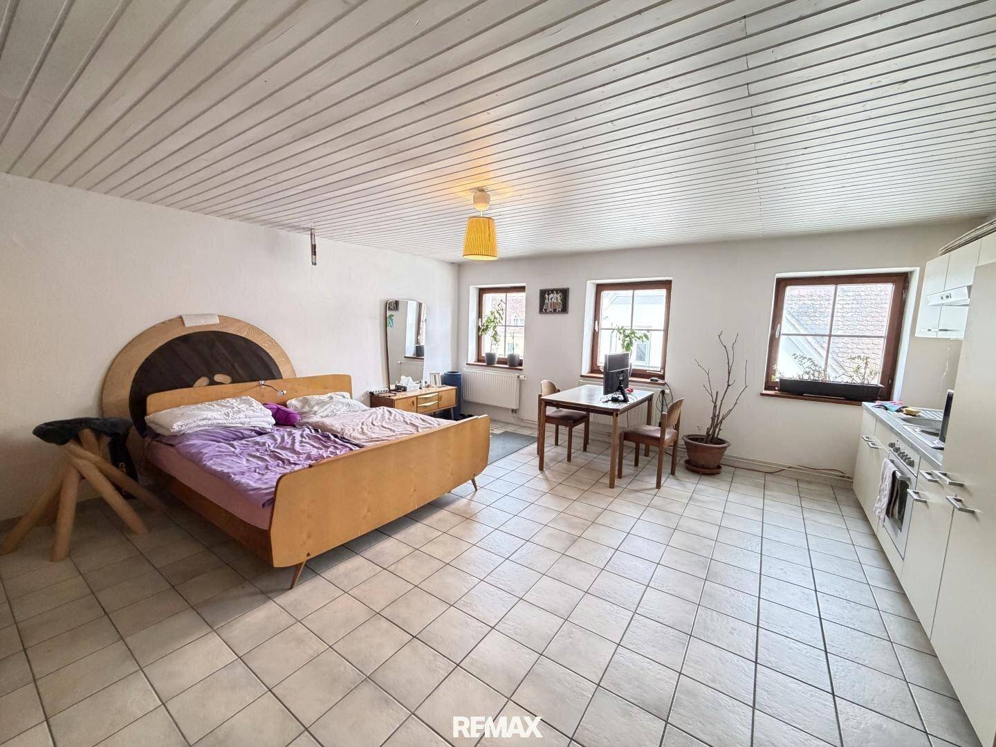 Geräumiges Zimmer mit Bett, Esstisch und integrierter Küchenzeile, gefliestem Boden und Holzdecke.