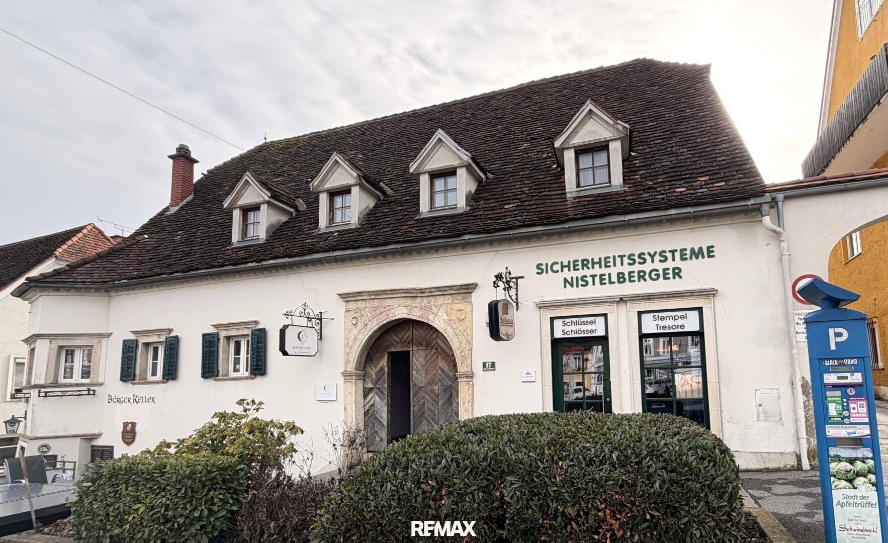 Ansicht der historischen Gebäudefassade mit Dachgauben, Geschäftsfront und einem Parkautomaten davor.