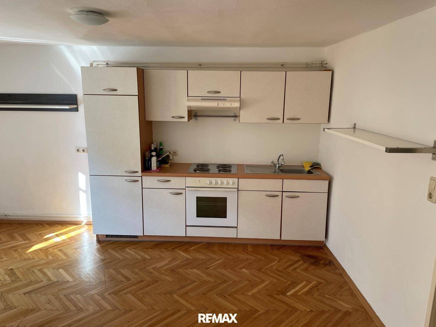 Einfache Küchenzeile mit weißen Schränken, Herd und Kühlschrank in einem Raum mit Parkettboden.