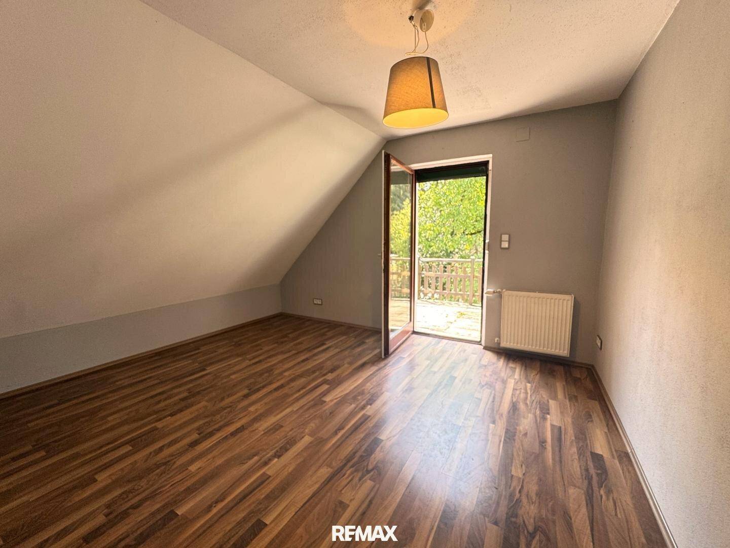 Leeres Zimmer mit Holzboden, Dachschräge und Zugang zu einem Balkon.