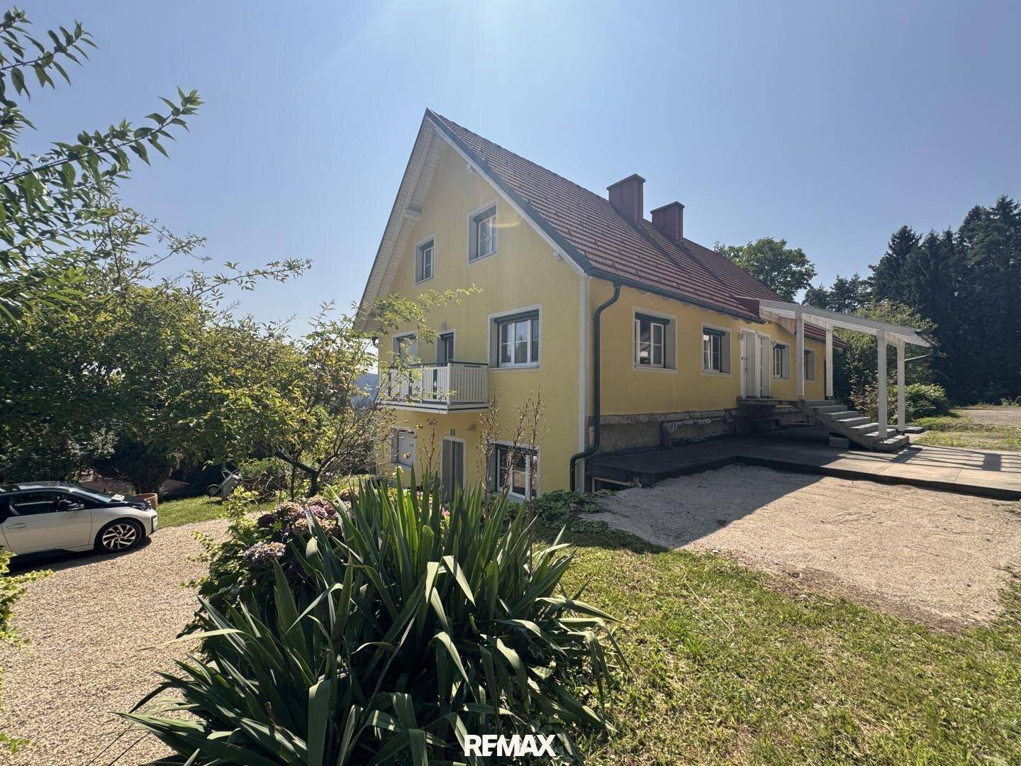 Gelbes Haus mit rotem Dach, Balkon und Kiesauffahrt an einem sonnigen Tag.