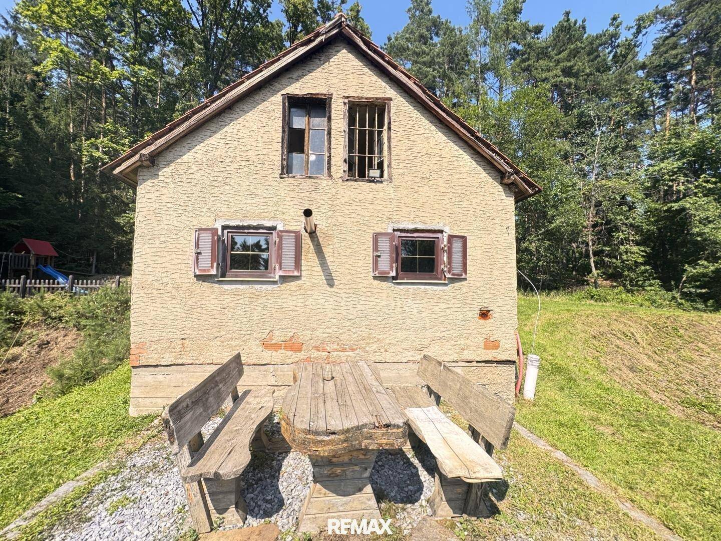 Rustikales Haus mit Holztisch und Bänken im Grünen, umgeben von Bäumen.