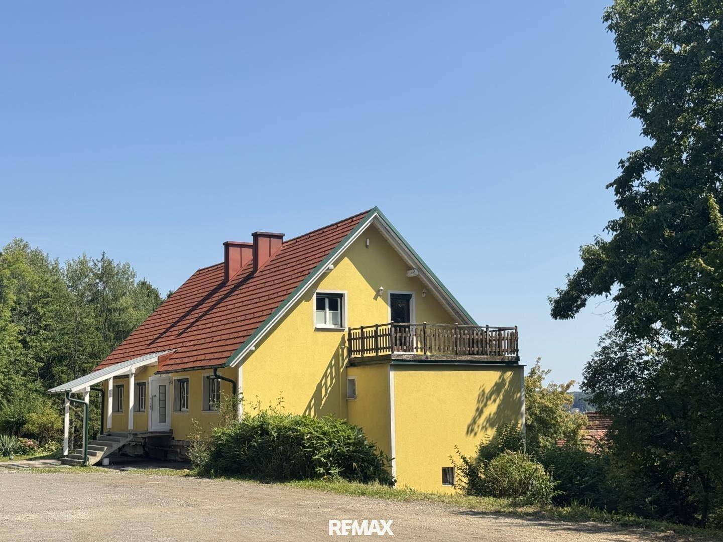 Großer Balkon mit Holzgeländer am gelben Haus, umgeben von Bäumen und mit Weitblick.