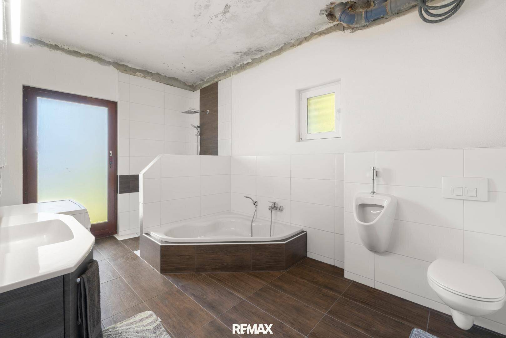 Badezimmer im Umbau mit Eckbadewanne, Urinal, Waschbecken und WC, teilweise gefliest und mit Fenster.