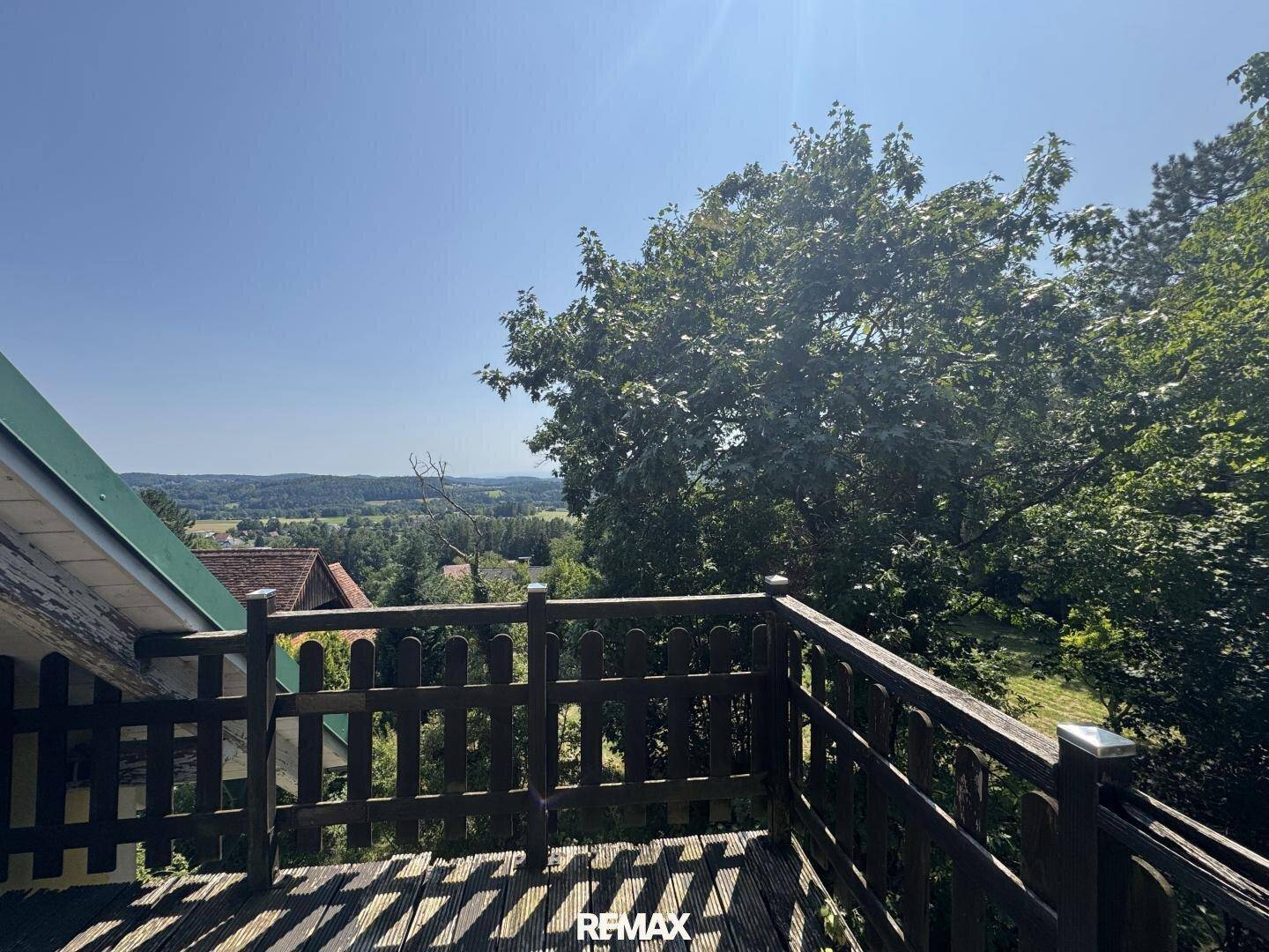 Holzbalkon mit Geländer und weitem Panoramablick über die grüne Landschaft unter blauem Himmel.