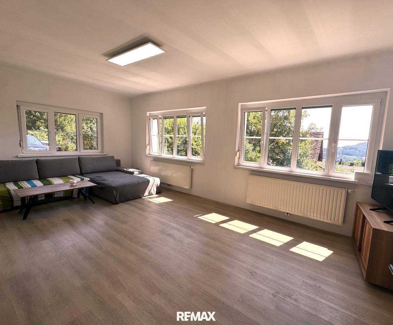 Helles Wohnzimmer mit bequemer Couch, vielen Fenstern und Blick ins Grüne an einem sonnigen Tag.
