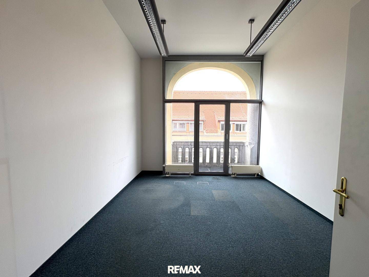 Leerer Raum mit dunklem Teppichboden und einem großen Bogenfenster, das viel Tageslicht hereinlässt.