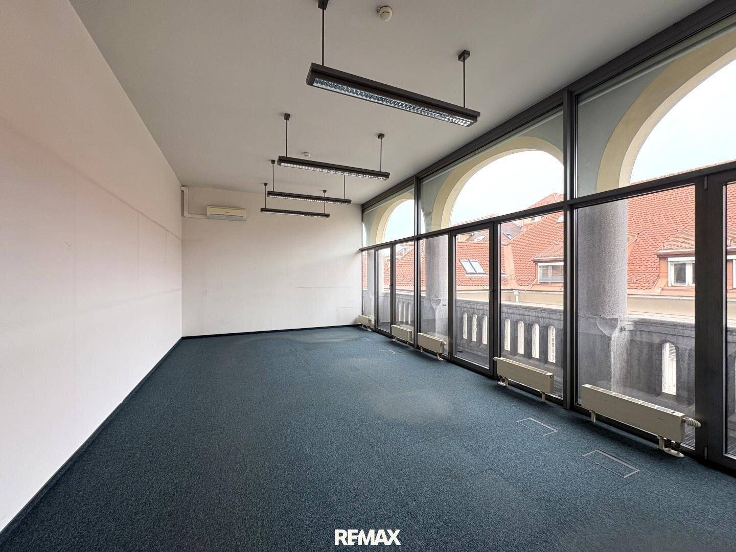 Geräumiger Büroraum mit dunklem Teppichboden, hohen Decken und großen Bogenfenstern mit Blick nach draußen.