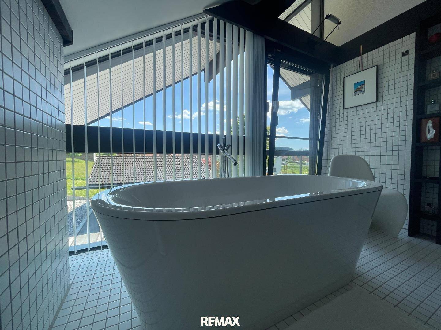 Freistehende Badewanne in einem hellen Badezimmer mit großen Fenstern und Blick ins Grüne.