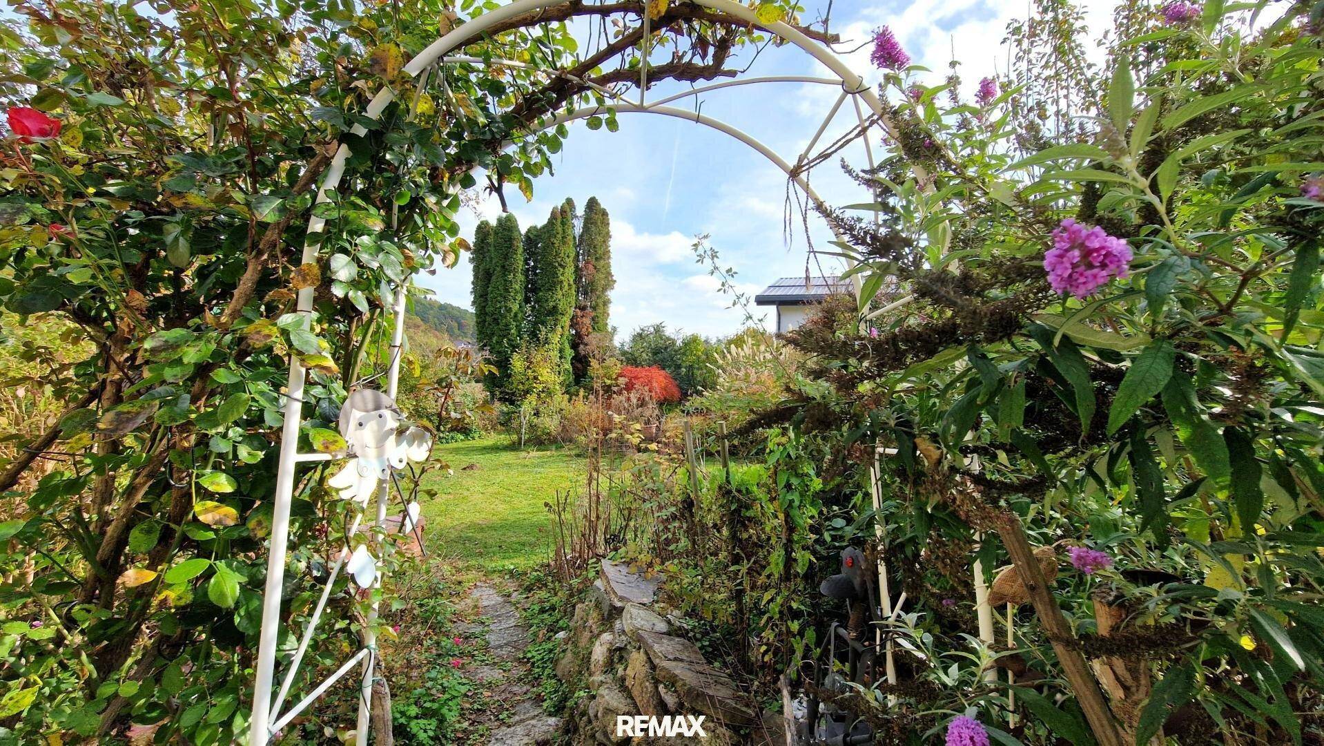 Romantischer Gartenweg unter einem Rosenbogen, der zu einer weitläufigen Grünfläche führt.