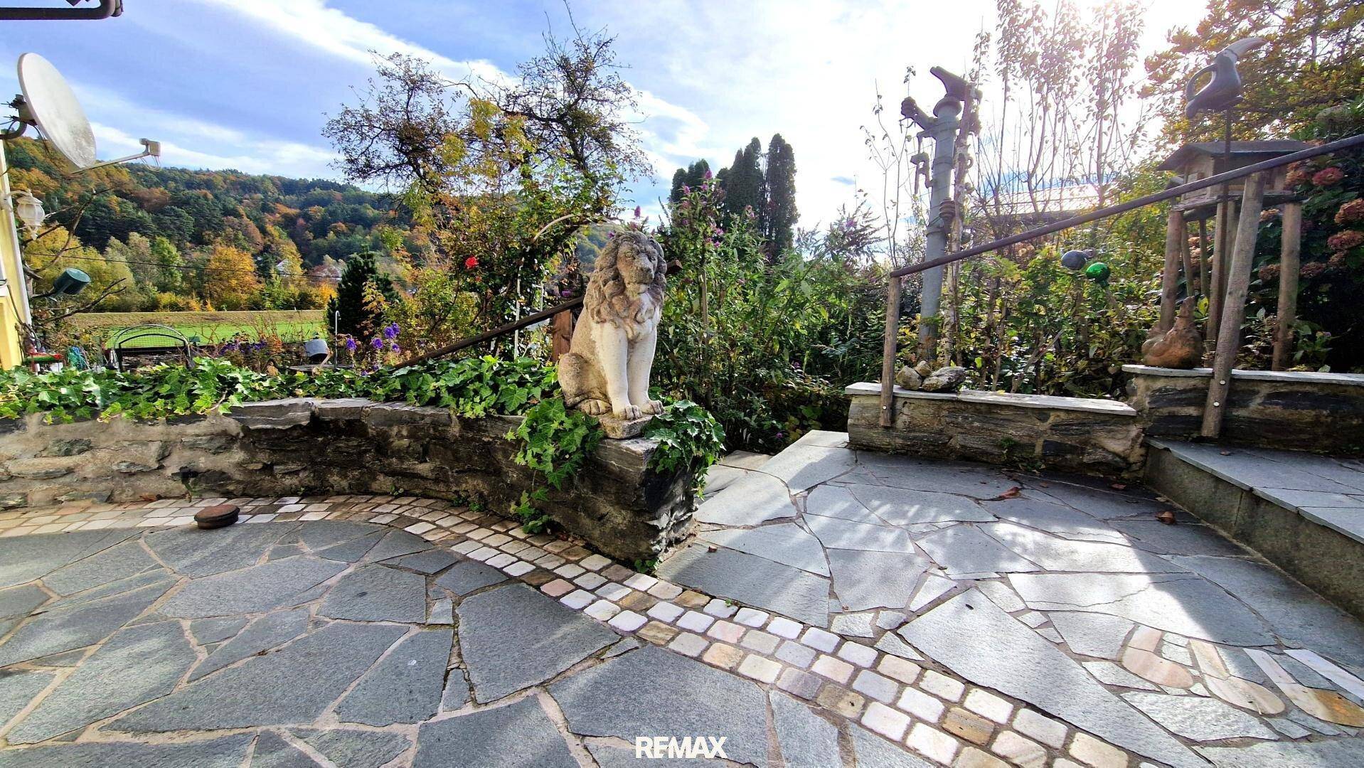 Gepflasterte Terrasse mit Steinmauern und einer Löwenstatue, umgeben von Grün.