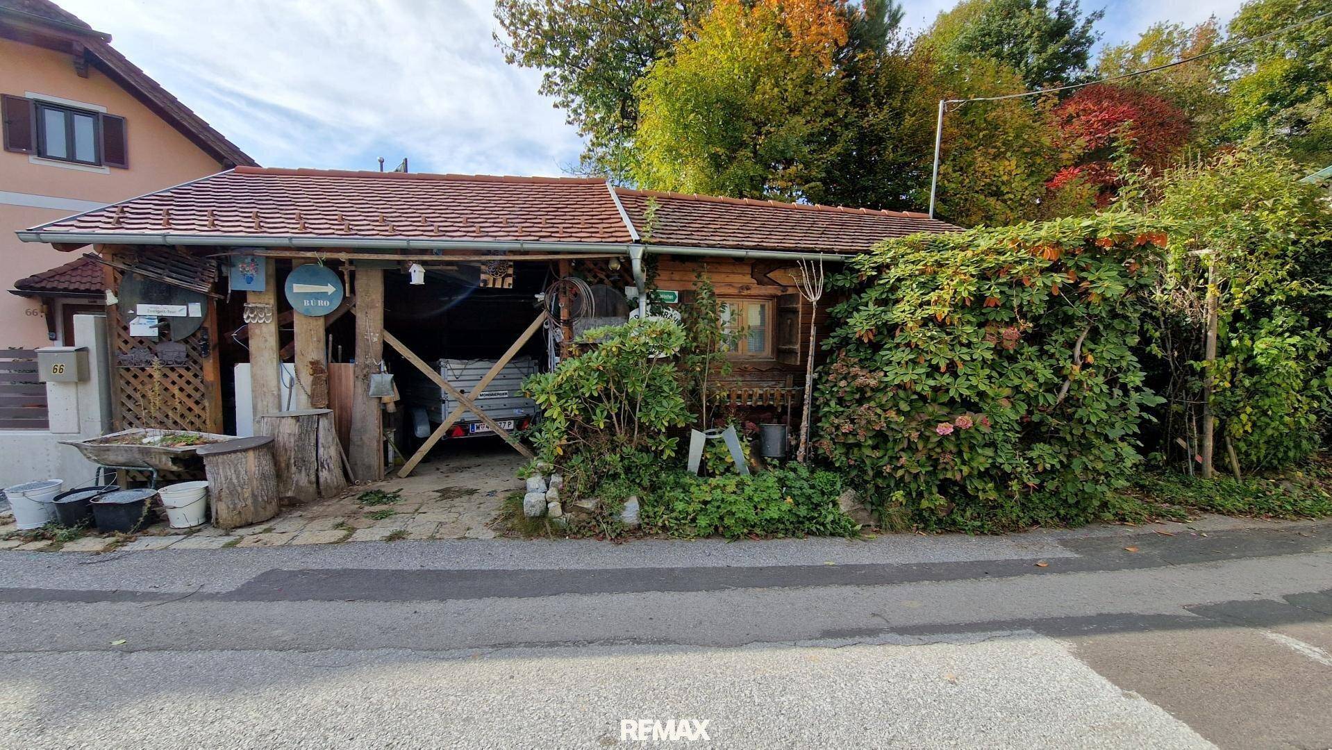 Rustikaler Holzunterstand oder Carport neben einer Straße, umgeben von Bäumen und einem Wohnhaus.