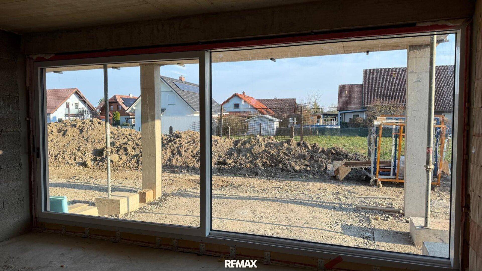 Innenraum im Rohbau mit großen Fenstern und Blick auf die Baustelle und Nachbarhäuser.