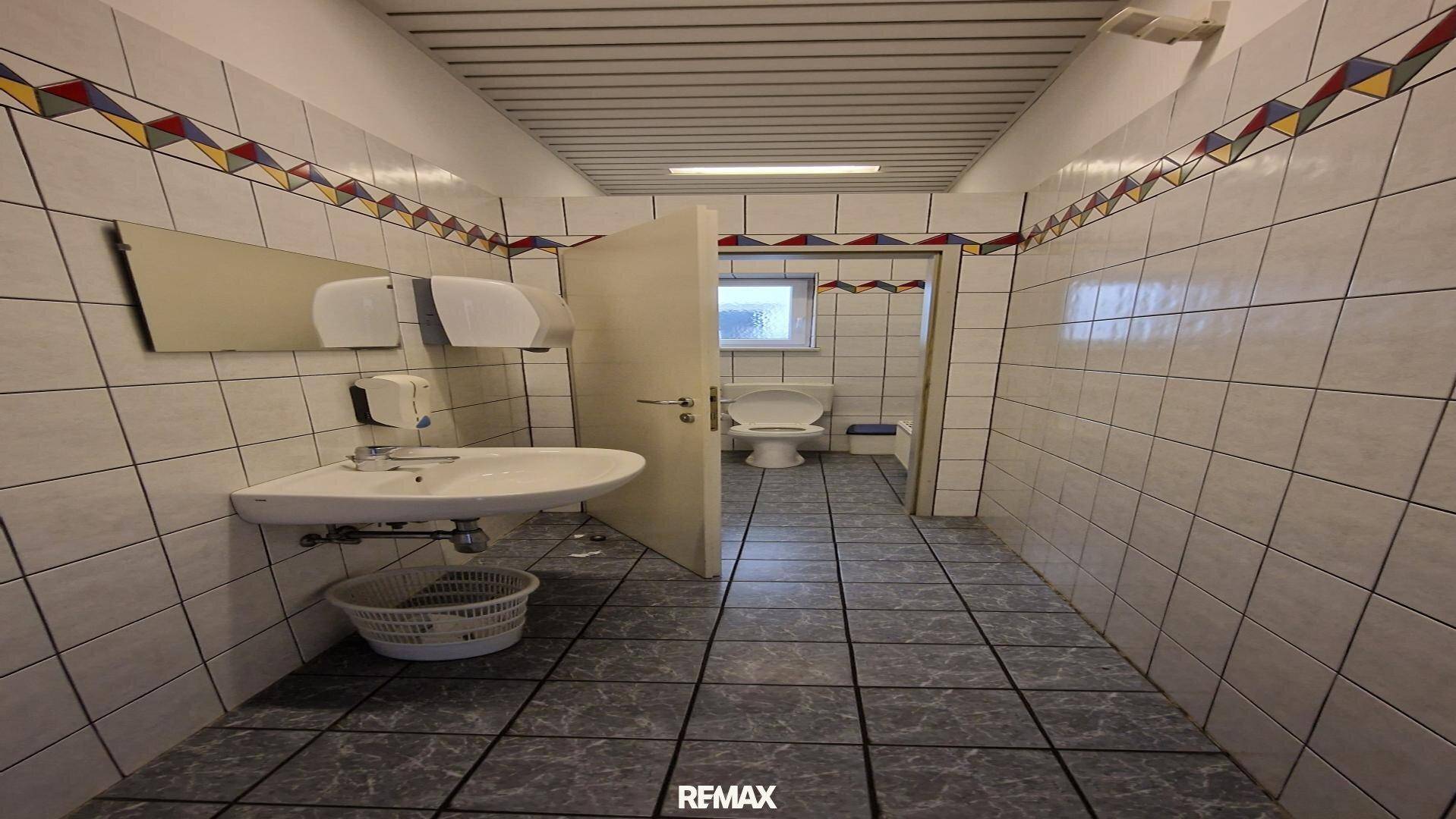 Geräumiger öffentlicher Waschraum mit weißen Fliesen, Waschbecken und Toilette in separatem Bereich, sauber und hell.