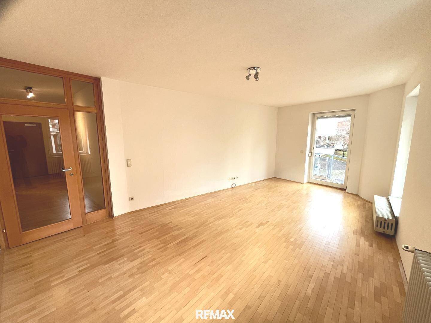 Geräumiges, leeres Wohnzimmer mit Parkettboden, weißen Wänden und einer Glastür zum Flur.