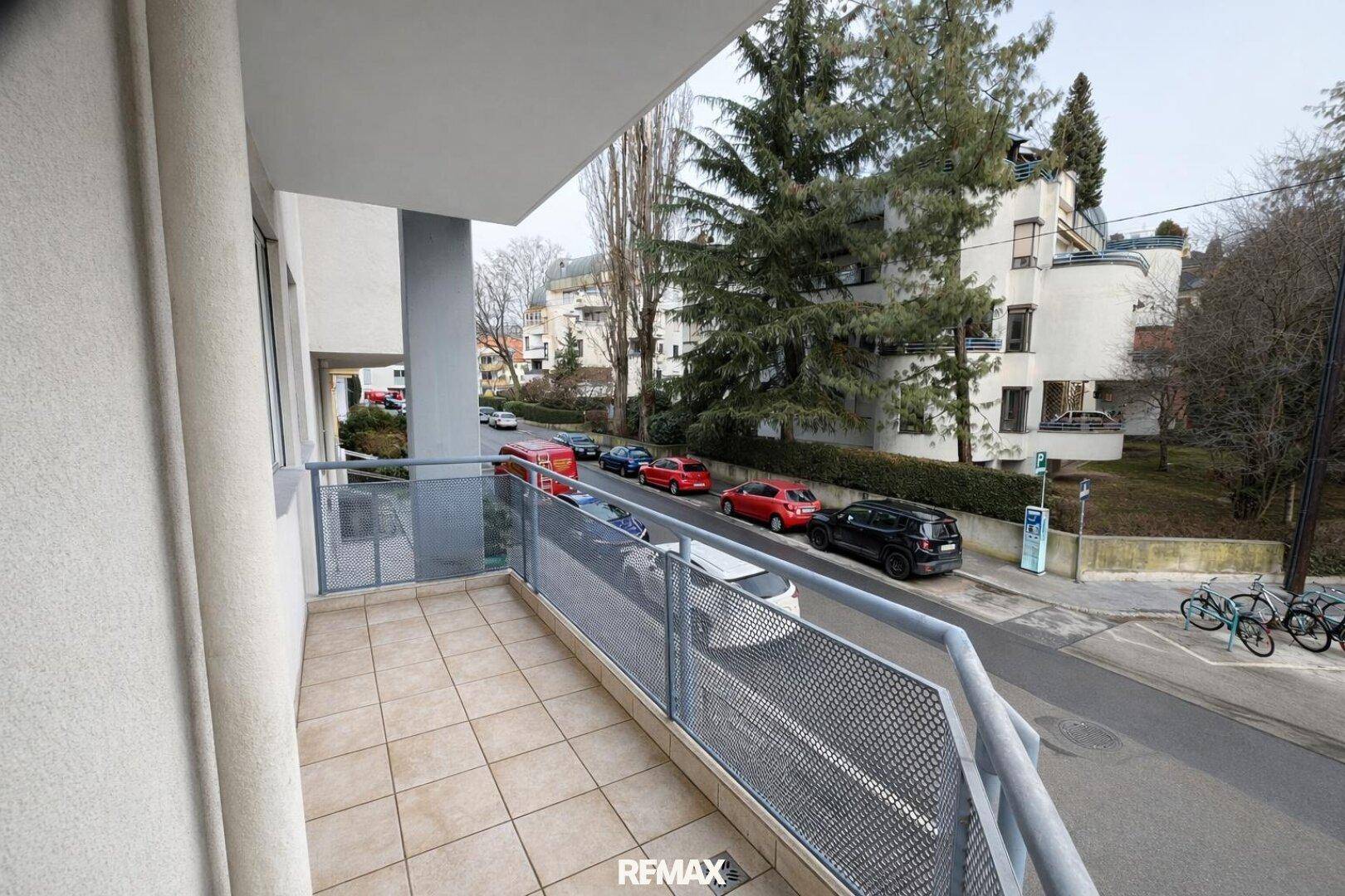 Leerer Balkon mit Fliesenboden und Metallgeländer, bietet Blick auf die Straße und Bäume.