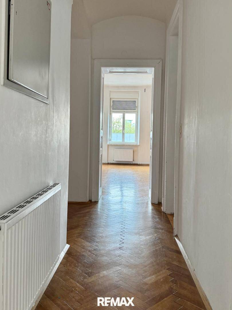 Heller Flur mit Fischgrätparkett und Heizkörper, der zu einem weiteren Raum mit Fenster führt.