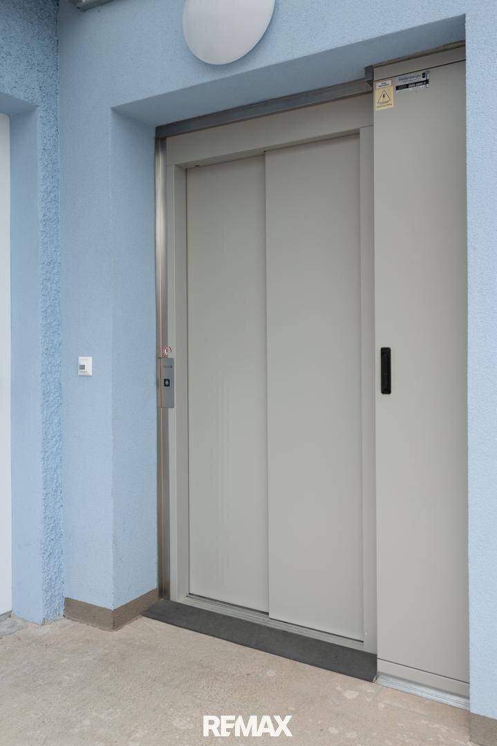 Geschlossener Aufzug in einem hellblauen Flur, mit Hinweisschildern und einem Griff.