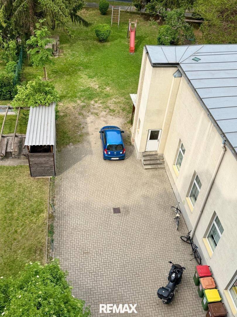 Luftaufnahme eines gepflasterten Parkplatzes neben einem Gebäude mit einem blauen Auto und Mülltonnen.