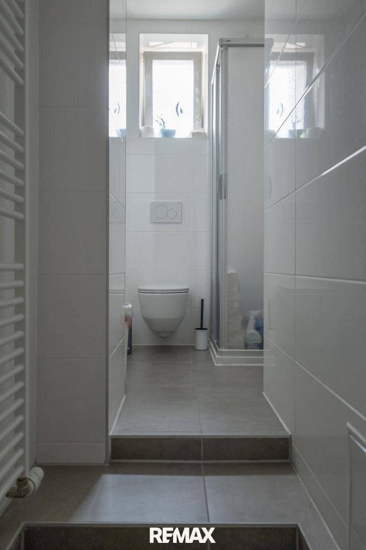 Schmaler, gefliester Badezimmerbereich mit wandhängendem WC und Duschkabine, hell beleuchtet.