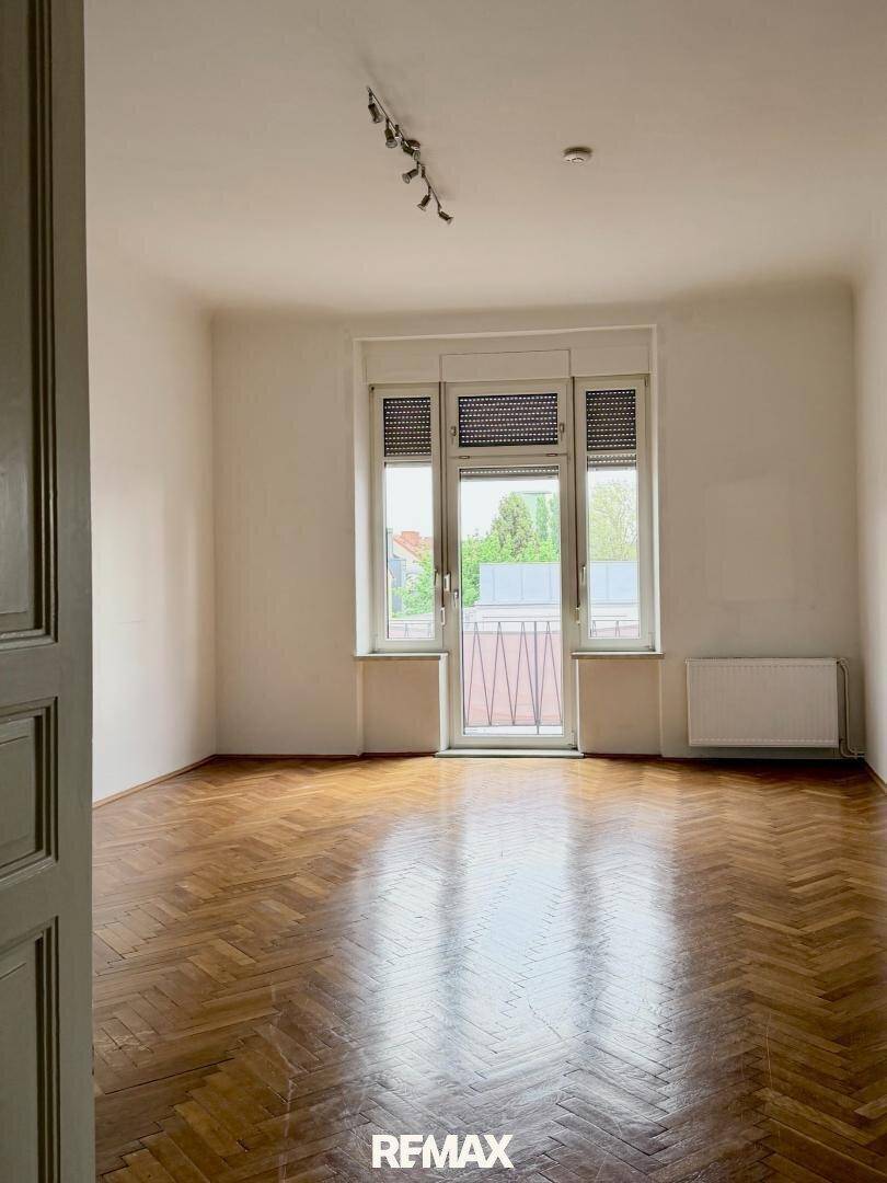 Heller, leerer Raum mit Fischgrätparkett und Zugang zu einem kleinen Balkon durch große Fenster.