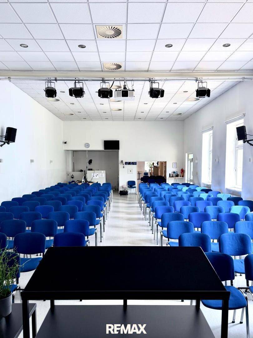 Geräumiger Konferenzsaal mit blauen Stuhlreihen, Projektor und einem schwarzen Präsentationstisch.
