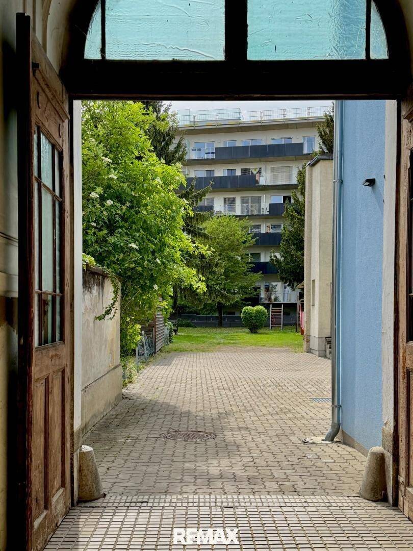 Blick durch eine offene Holztür auf einen gepflasterten Innenhof mit Grünflächen und einem Mehrfamilienhaus im Hintergrund.