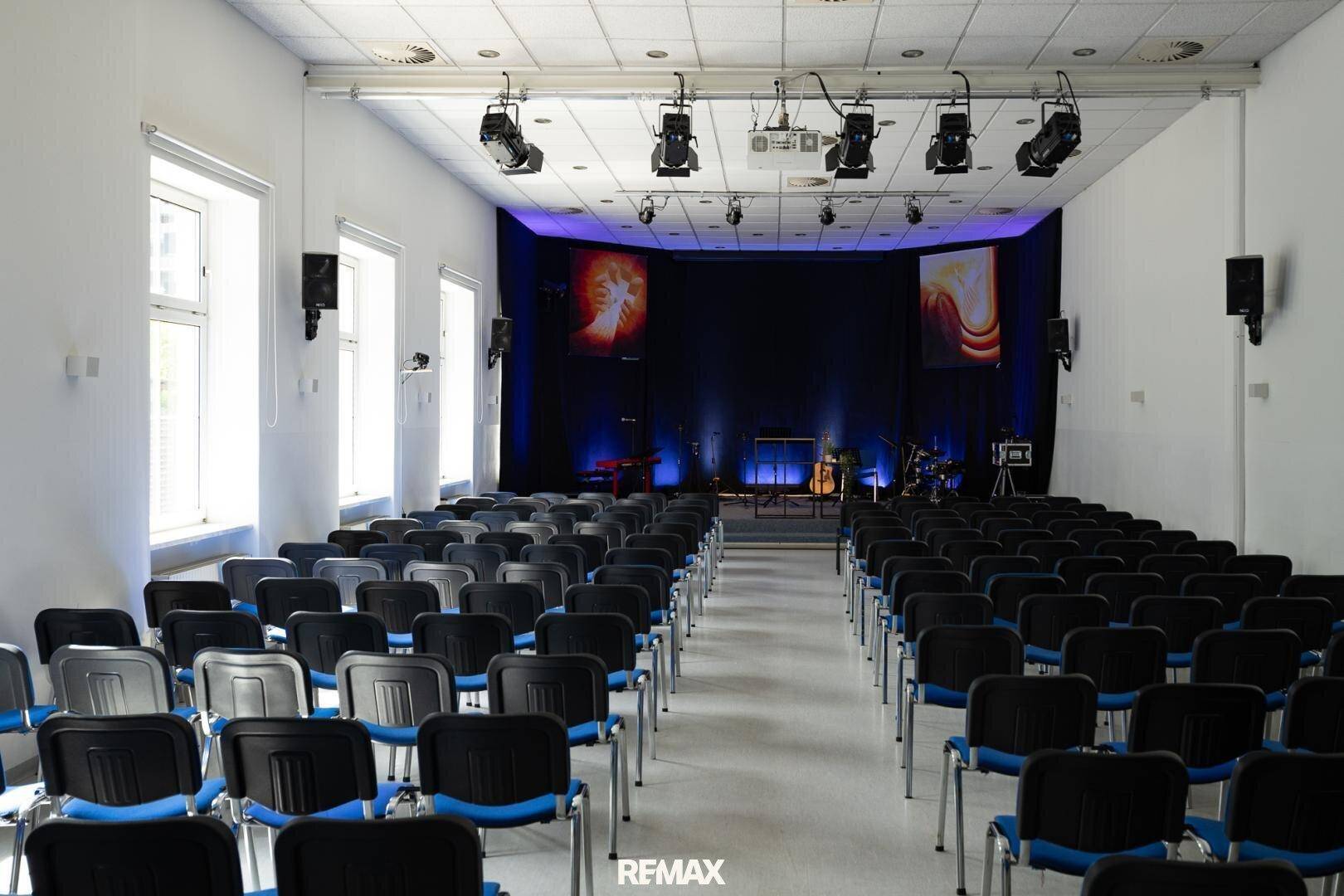 Großer Konferenzraum mit vielen Stühlen, Bühne und professioneller Beleuchtungs- und Soundanlage.