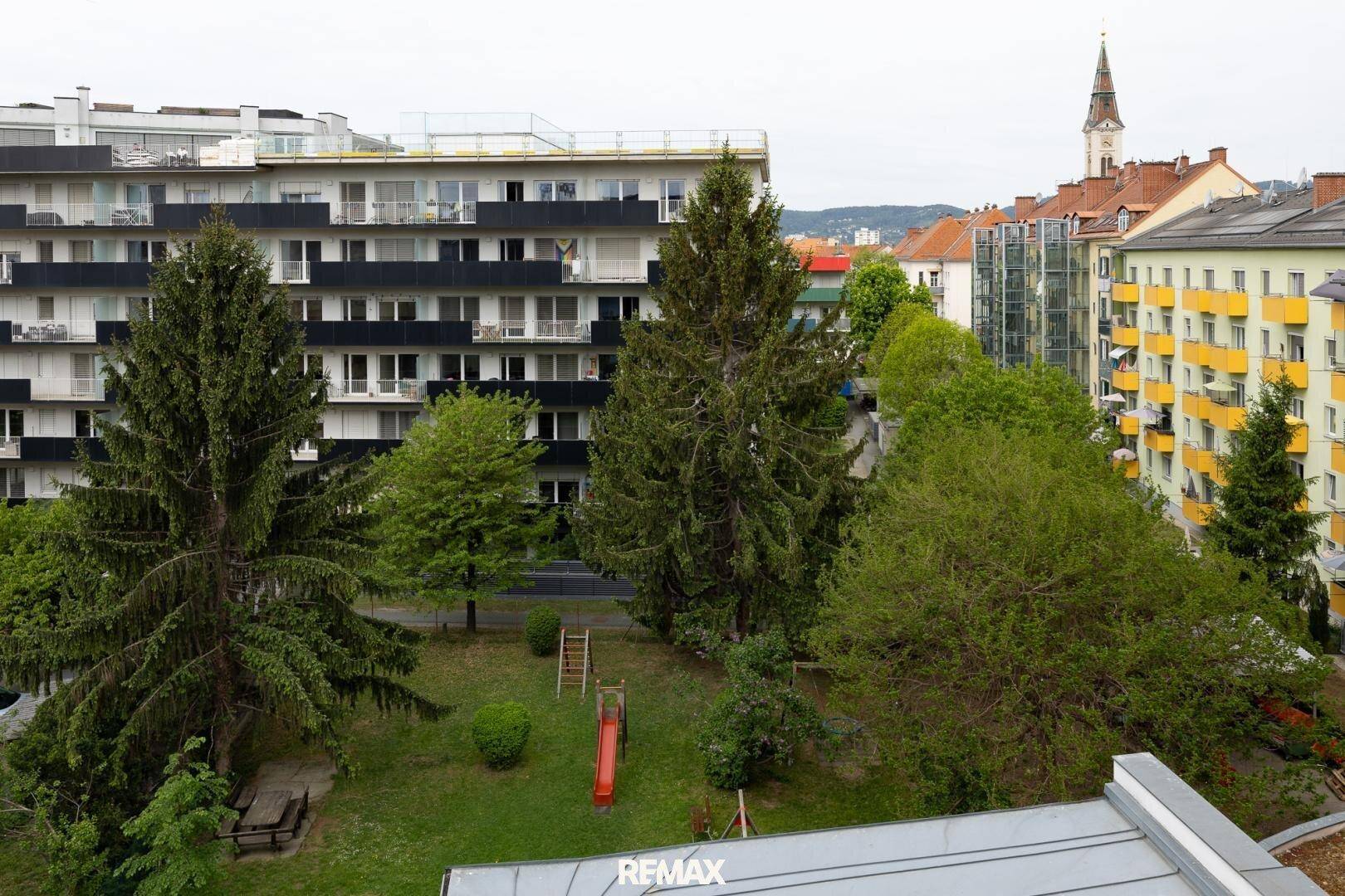 Panoramablick über eine Wohngegend mit mehreren Gebäuden, grünen Bäumen und einem Kirchturm in der Ferne.