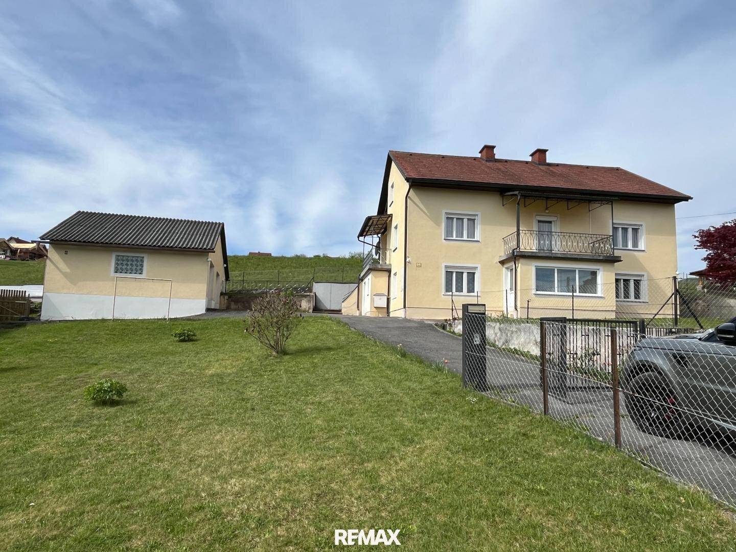 Gelbes Einfamilienhaus mit großem Garten und Nebengebäude unter blauem Himmel.