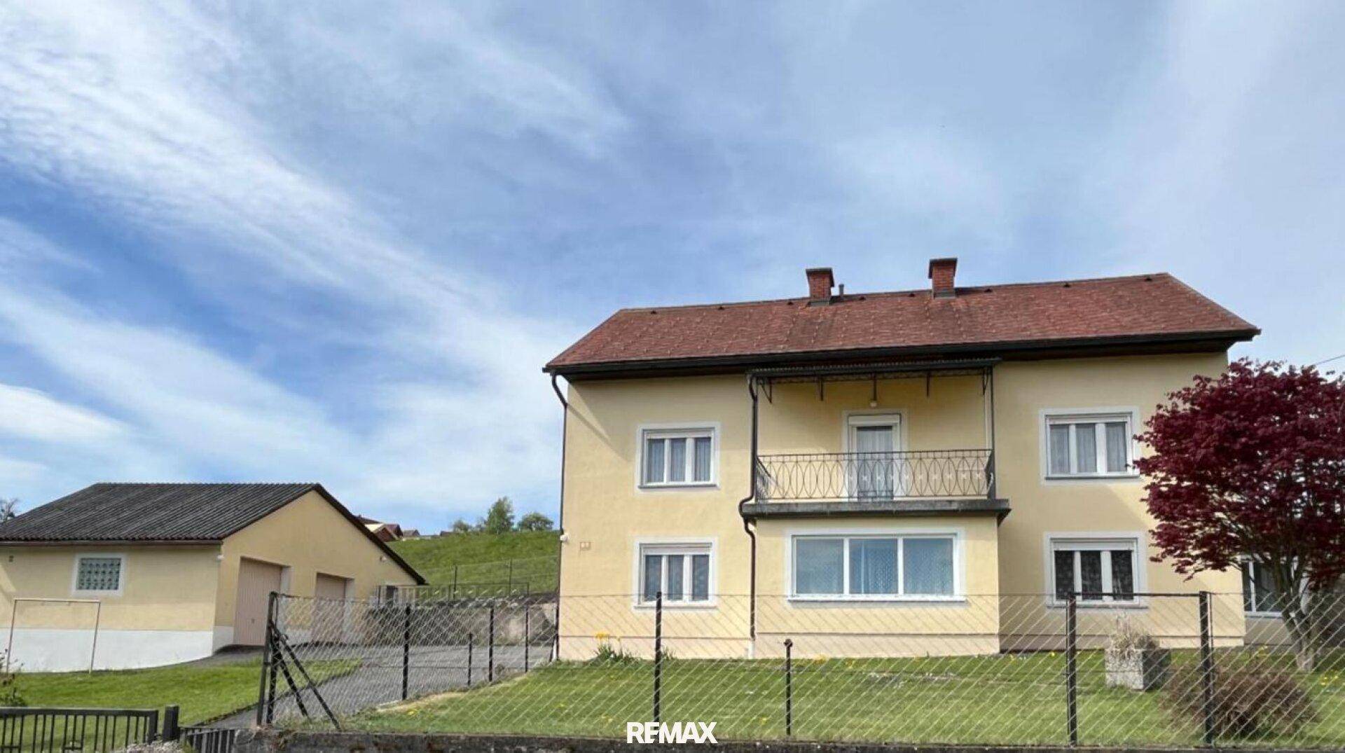 Gelbes Einfamilienhaus mit Garten und Nebengebäude unter blauem Himmel.