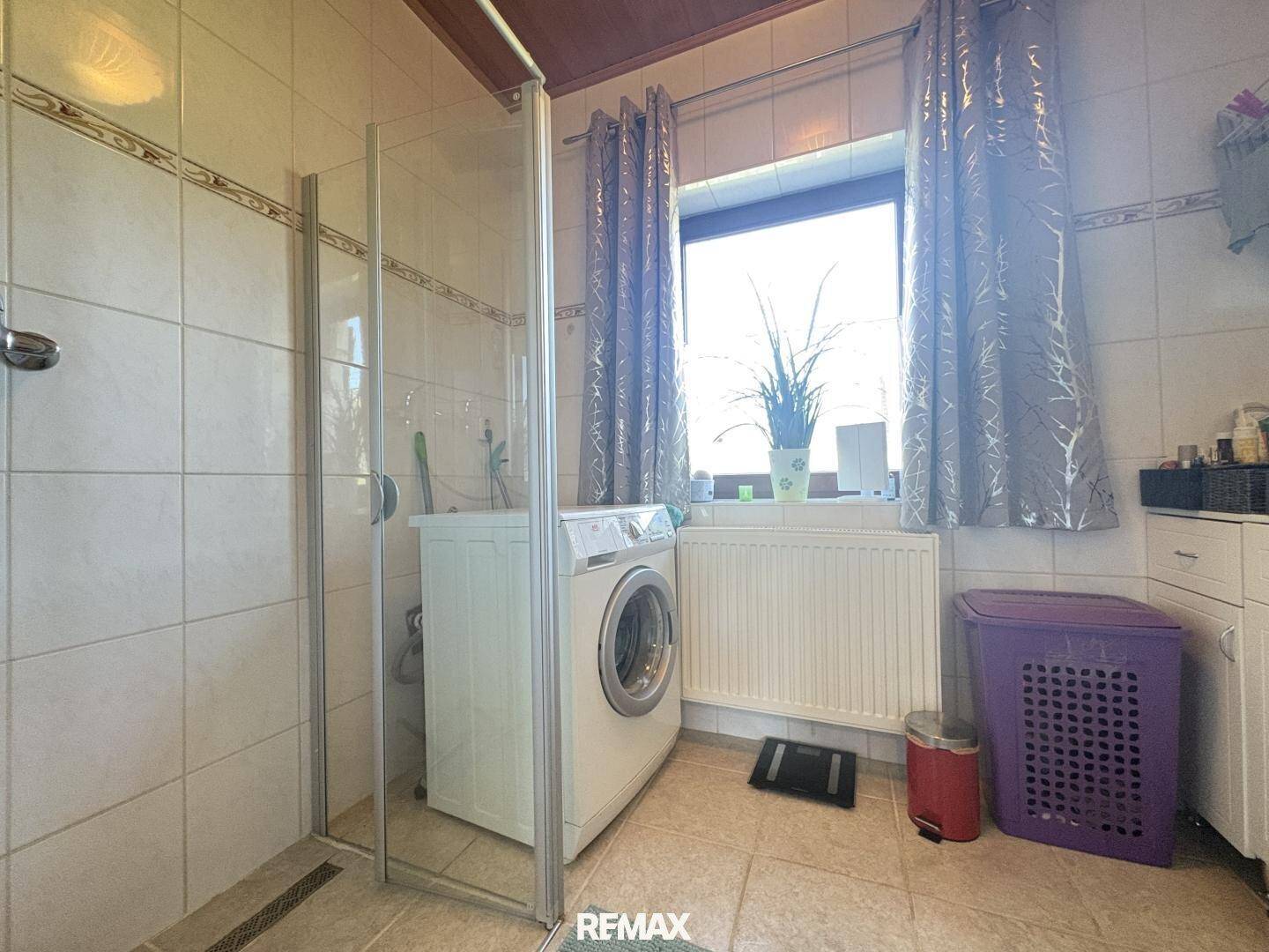 Badezimmer mit Duschkabine, Waschmaschine und Fenster, ausgestattet mit hellen Fliesen.