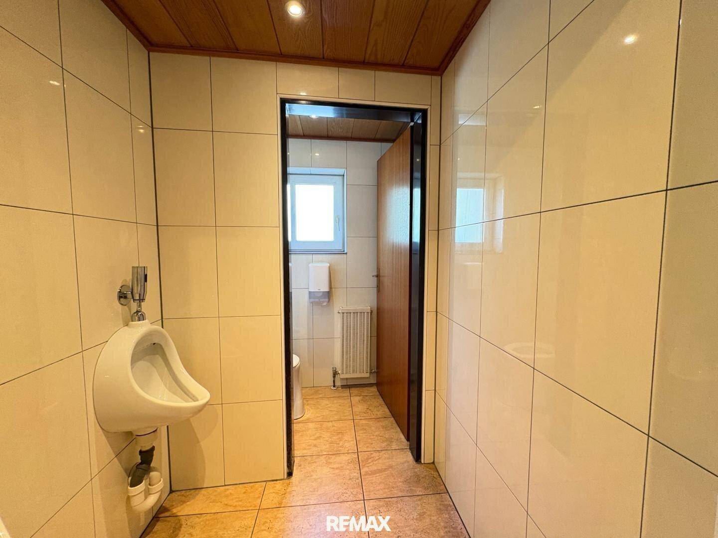 Gefliester WC-Bereich mit Urinal und Zugang zu weiteren Toiletten, hell und gepflegt.