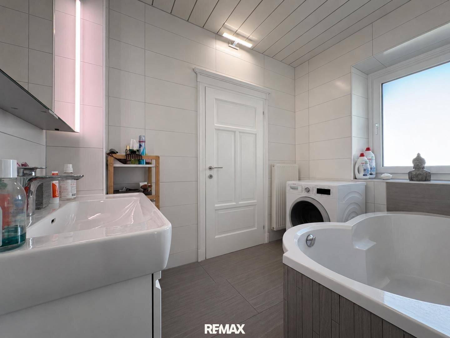Modern gefliestes Badezimmer mit Badewanne, Waschbecken und Waschmaschine, hell und funktional.